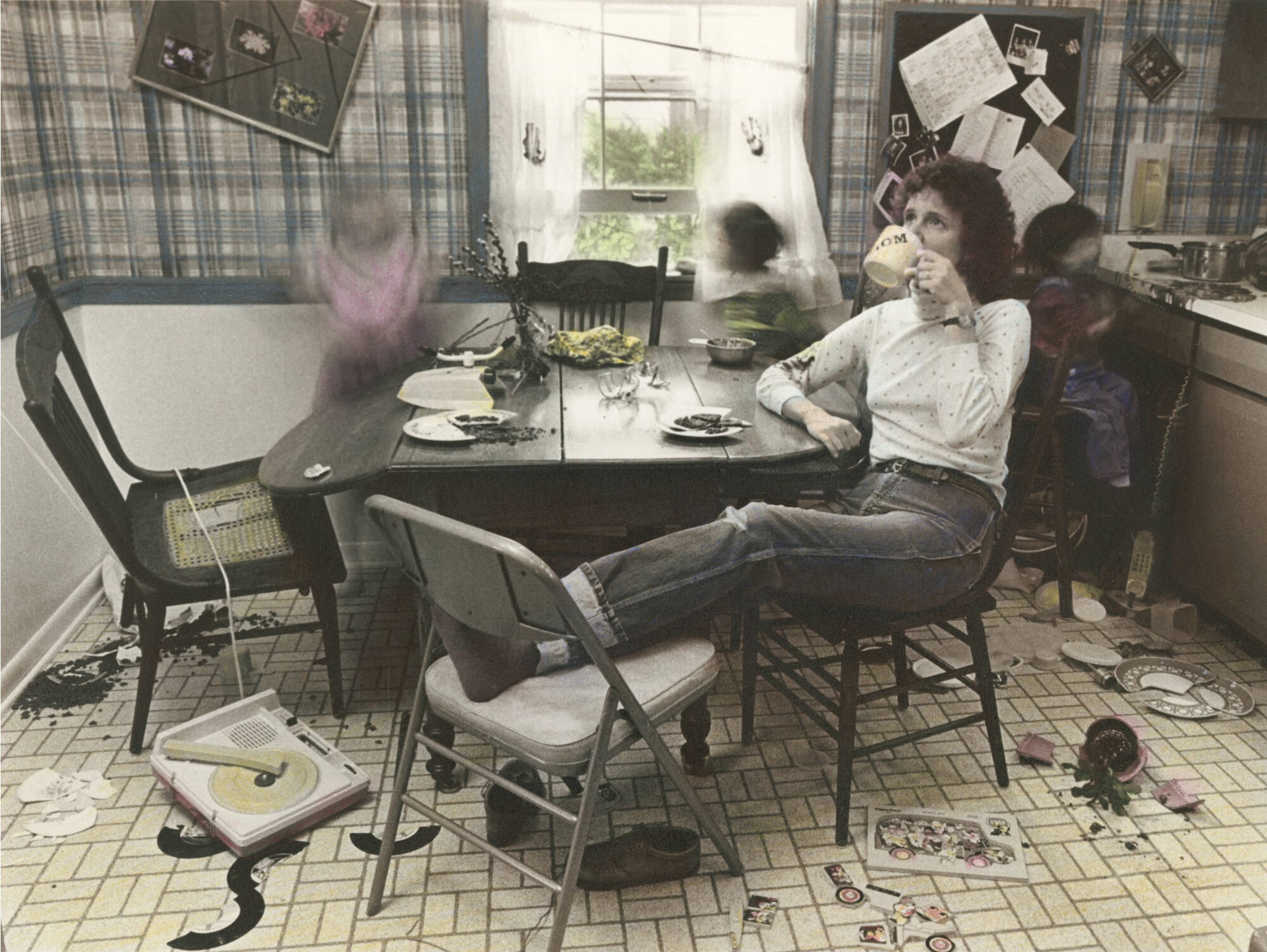 A hand-colored photographic print of a woman sitting at a kitchen table surrounded by chaos. She is drinking out of a coffee cup that says "mom".